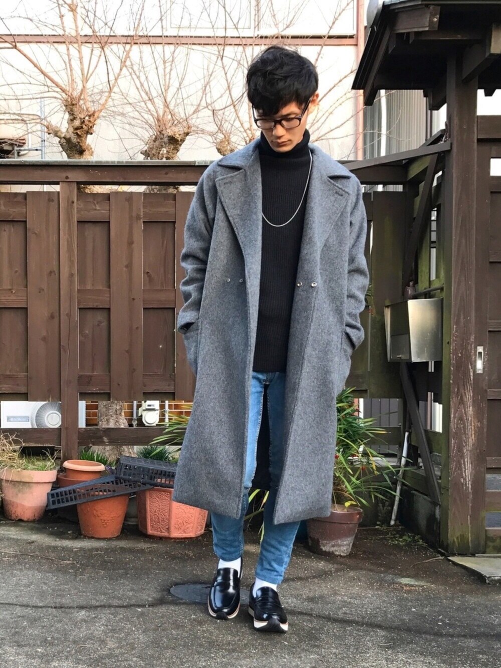 グレー系のチェスターコート、ブラック系のニット/セーター、ブルー系のデニムパンツを着用したメンズの冬コーディネートの1枚目の写真