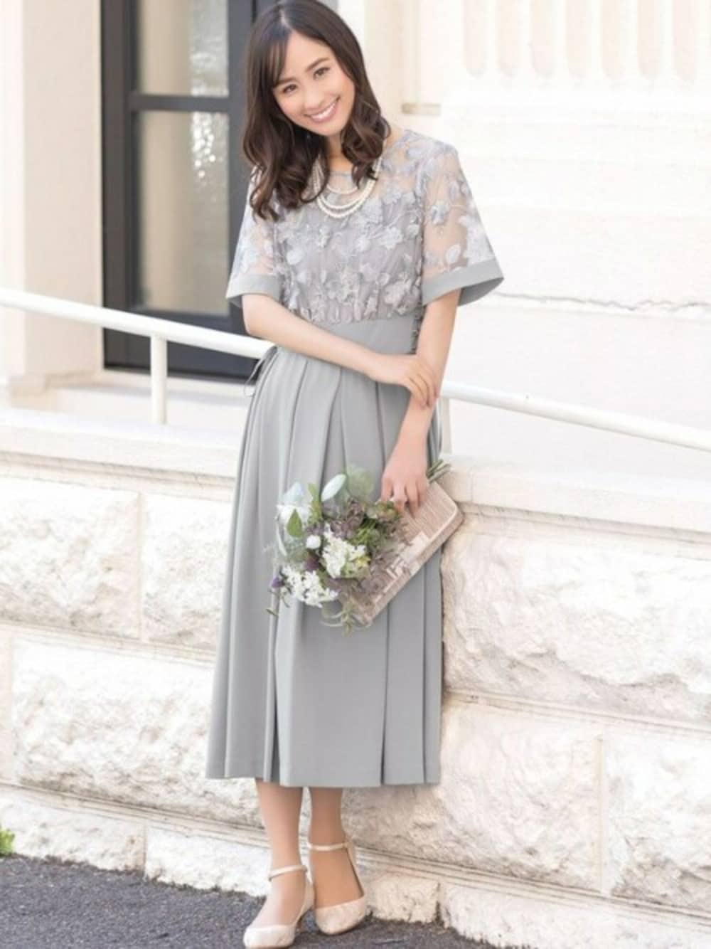 グレー系のドレス、ベージュ系のクラッチバッグ、ベージュ系のパンプスを着用したレディースの春コーディネートの1枚目の写真