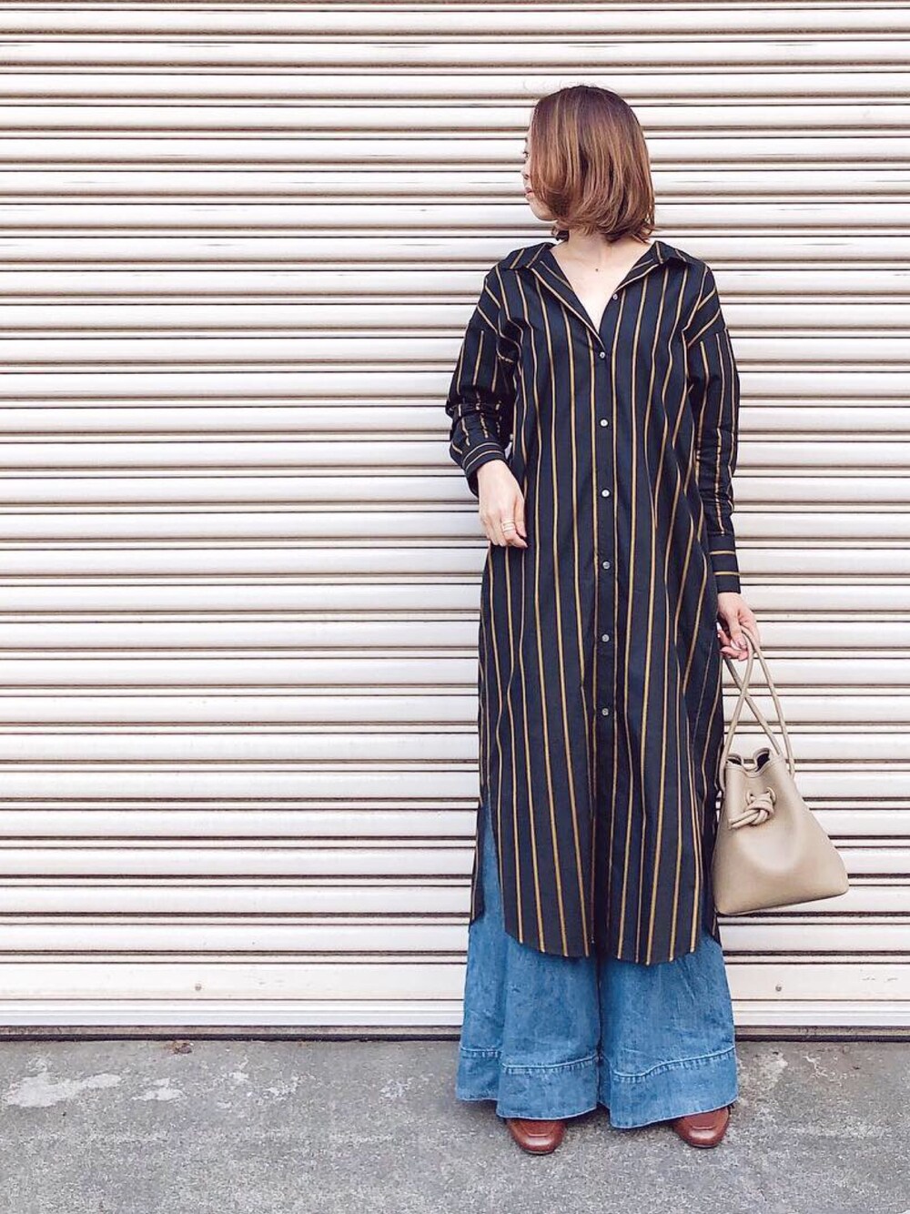 ブラック系のシャツワンピース、ベージュ系のハンドバッグ、ブルー系のデニムパンツを着用したレディースの秋コーディネートの1枚目の写真