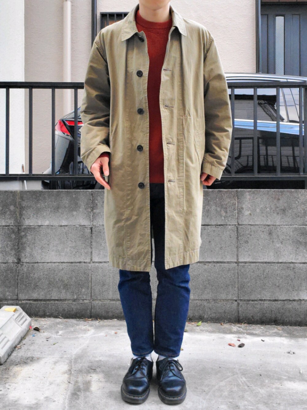 グリーン系のステンカラーコート、オレンジ系のニット/セーター、ブルー系のデニムパンツを着用したメンズの春コーディネートの1枚目の写真