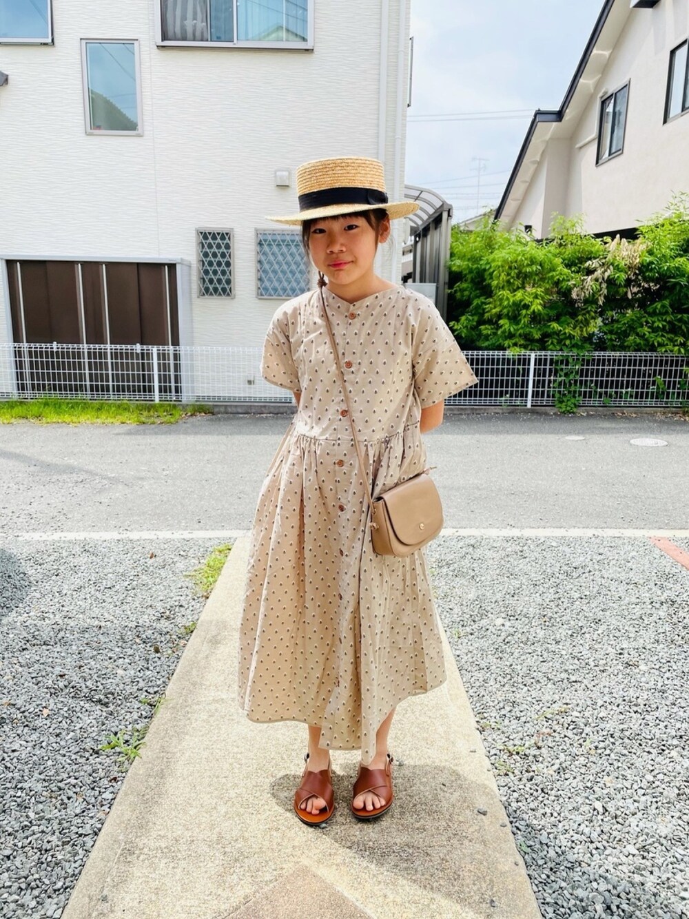 ベージュ系のショルダーバッグ、ベージュ系のシャツワンピース、ブラウン系のサンダルを着用したキッズの夏コーディネートの1枚目の写真