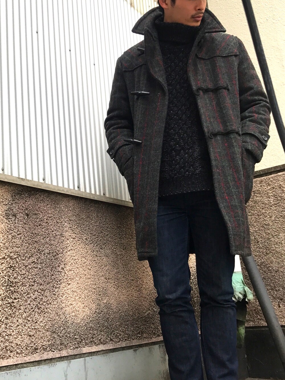 グレー系のダッフルコート、ブラック系のニット/セーター、ブルー系のデニムパンツを着用したメンズの冬コーディネートの1枚目の写真