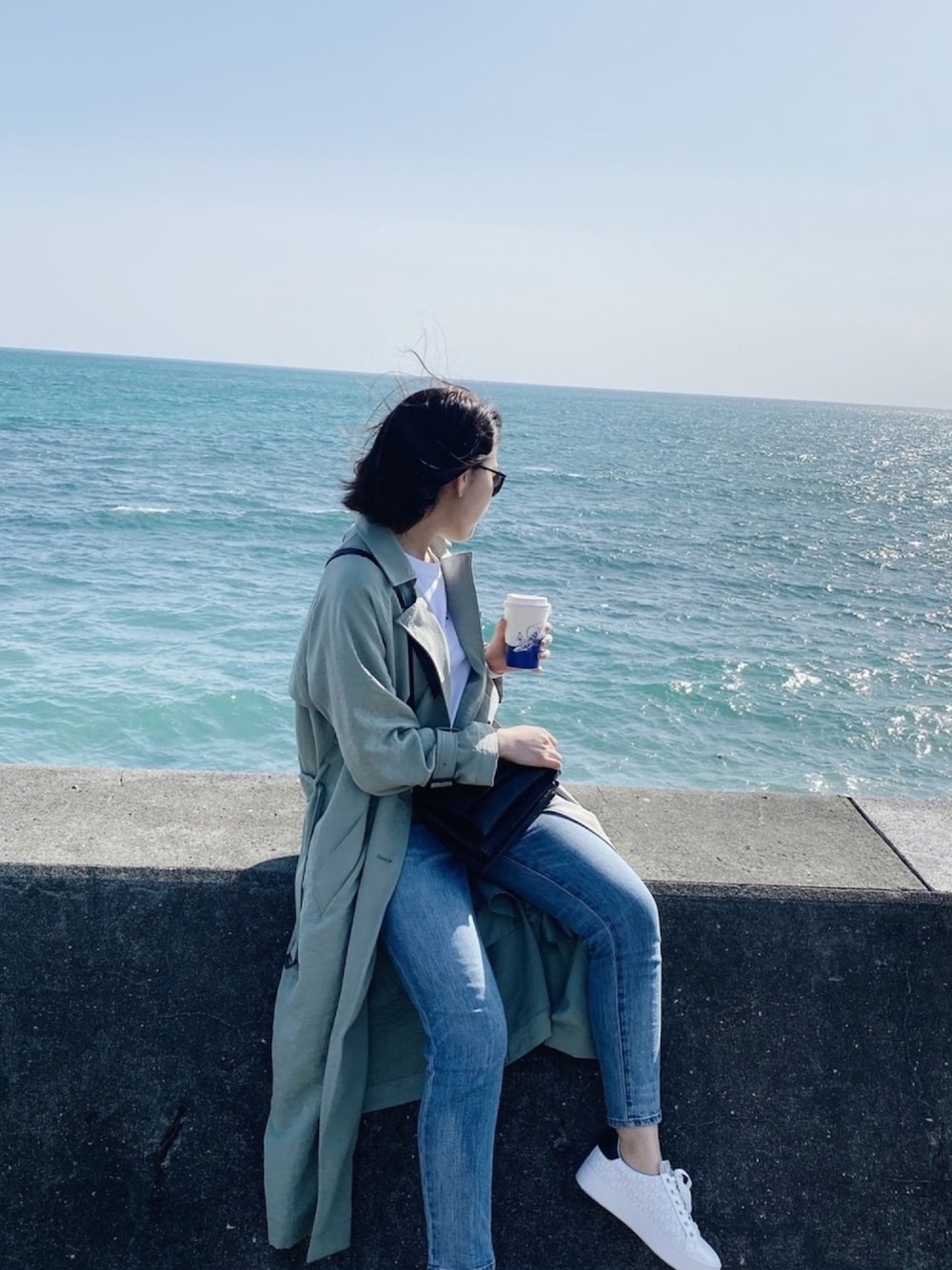 グリーン系のトレンチコート、ブラック系のデニムパンツ、ホワイト系のスニーカーを着用したレディースの春コーディネートの1枚目の写真