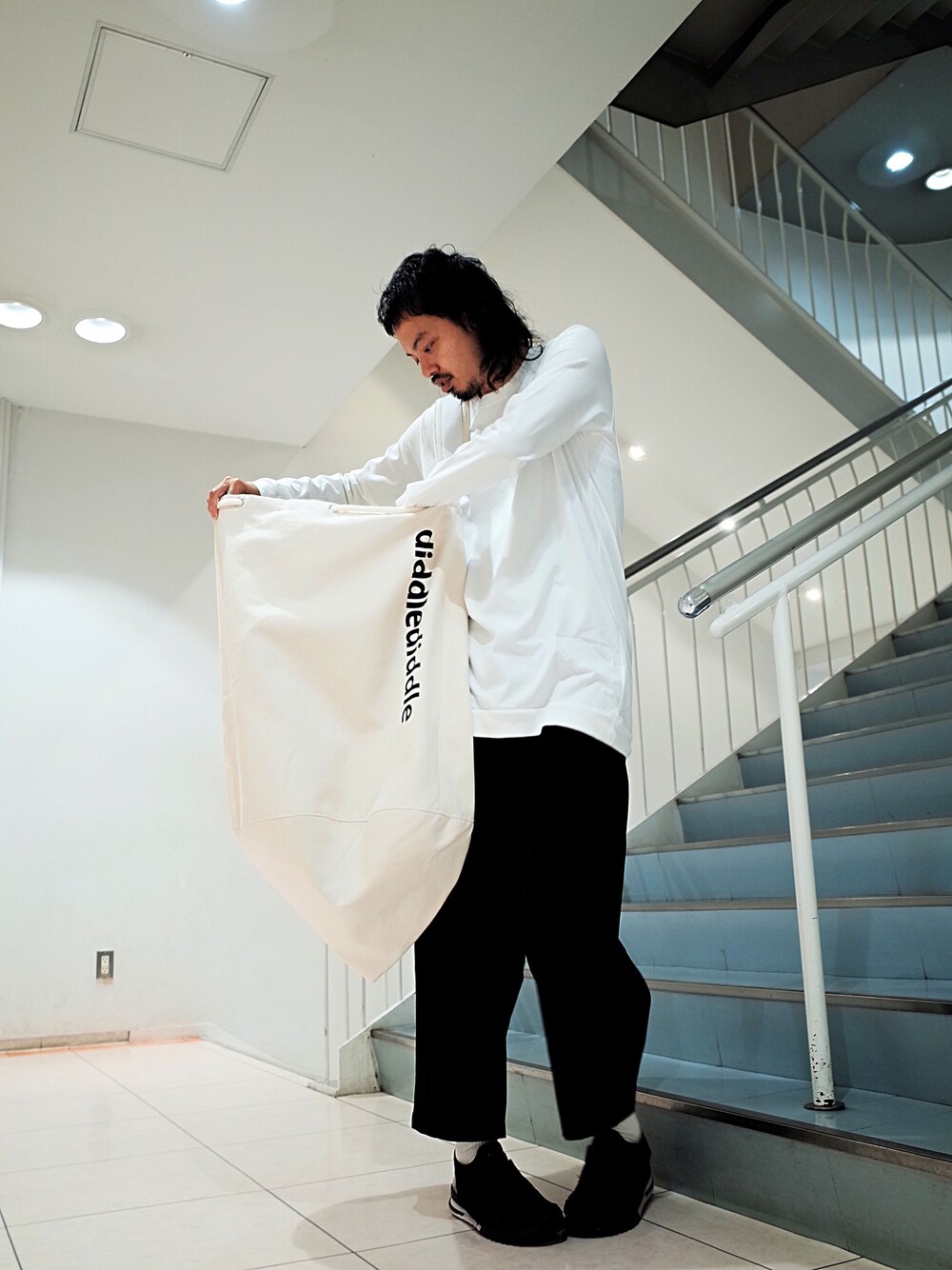 ホワイト系のTシャツ/カットソー、ホワイト系のトートバッグ、ブラック系のチノパンツを着用したメンズの夏コーディネートの1枚目の写真