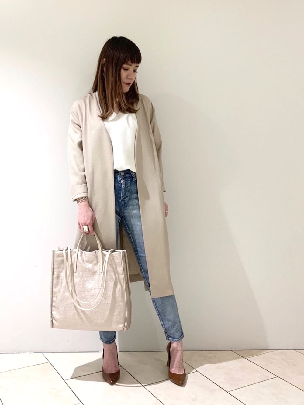 ベージュ系のノーカラーコート、ホワイト系のニット/セーター、ゴールド系のトートバッグを着用したレディースの春コーディネートの1枚目の写真