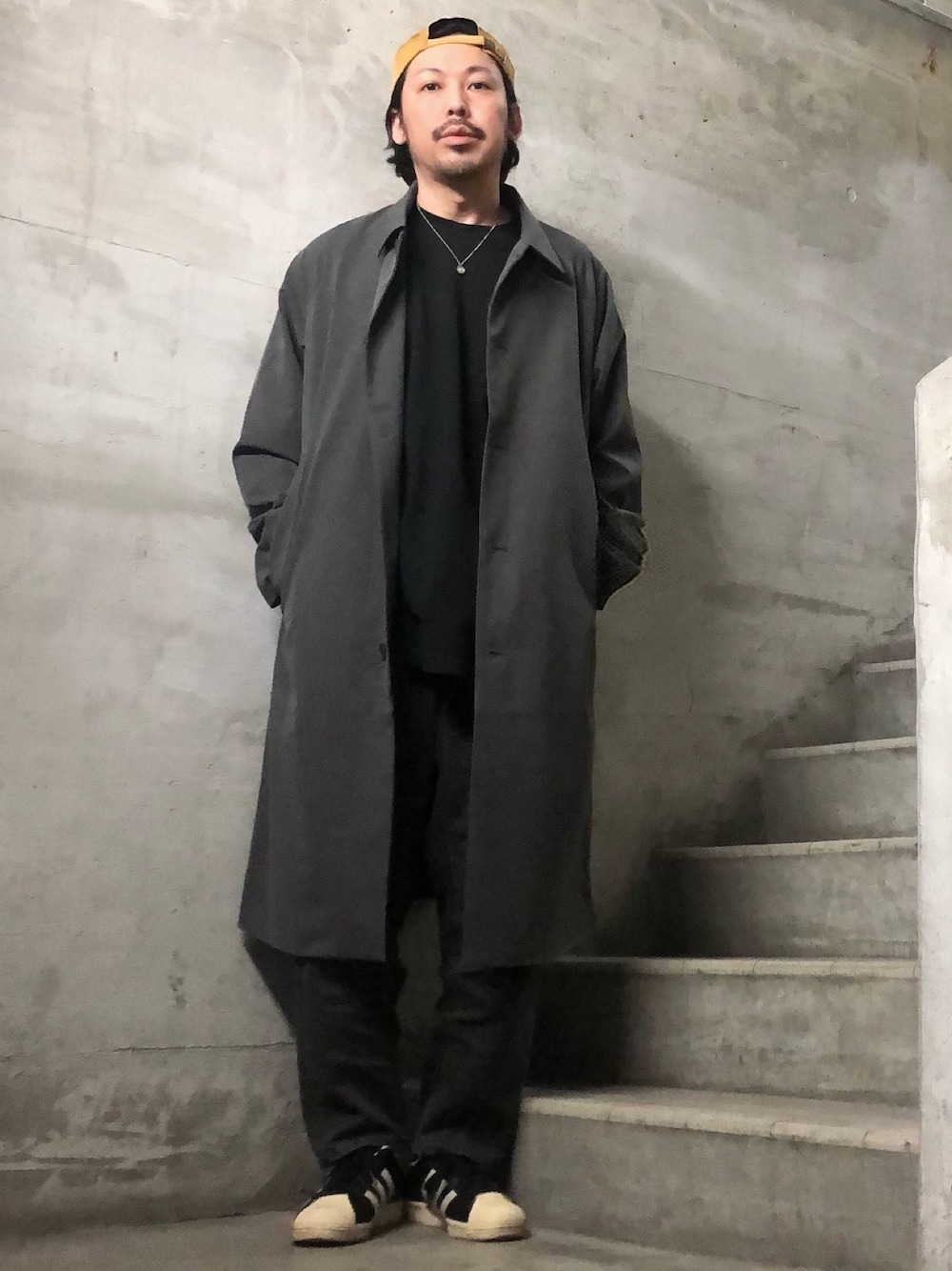 グレー系のステンカラーコート、ブラック系のスニーカー、ブラック系のTシャツ/カットソーを着用したメンズの冬コーディネートの1枚目の写真