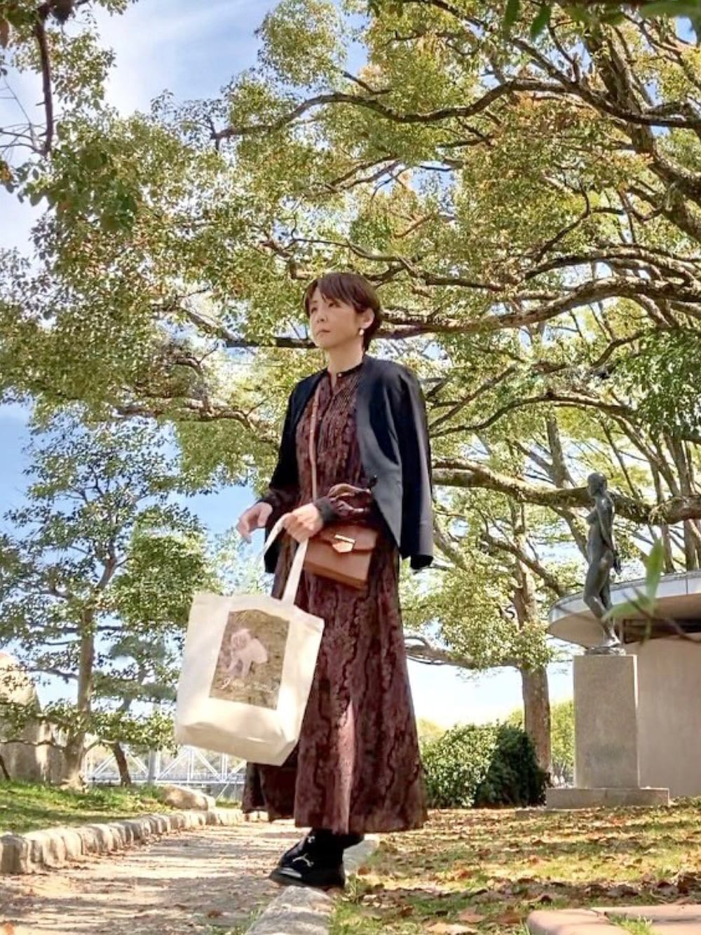 ブラック系のワンピース、ブラウン系のハンドバッグ、ベージュ系のトートバッグを着用したレディースの春コーディネートの1枚目の写真