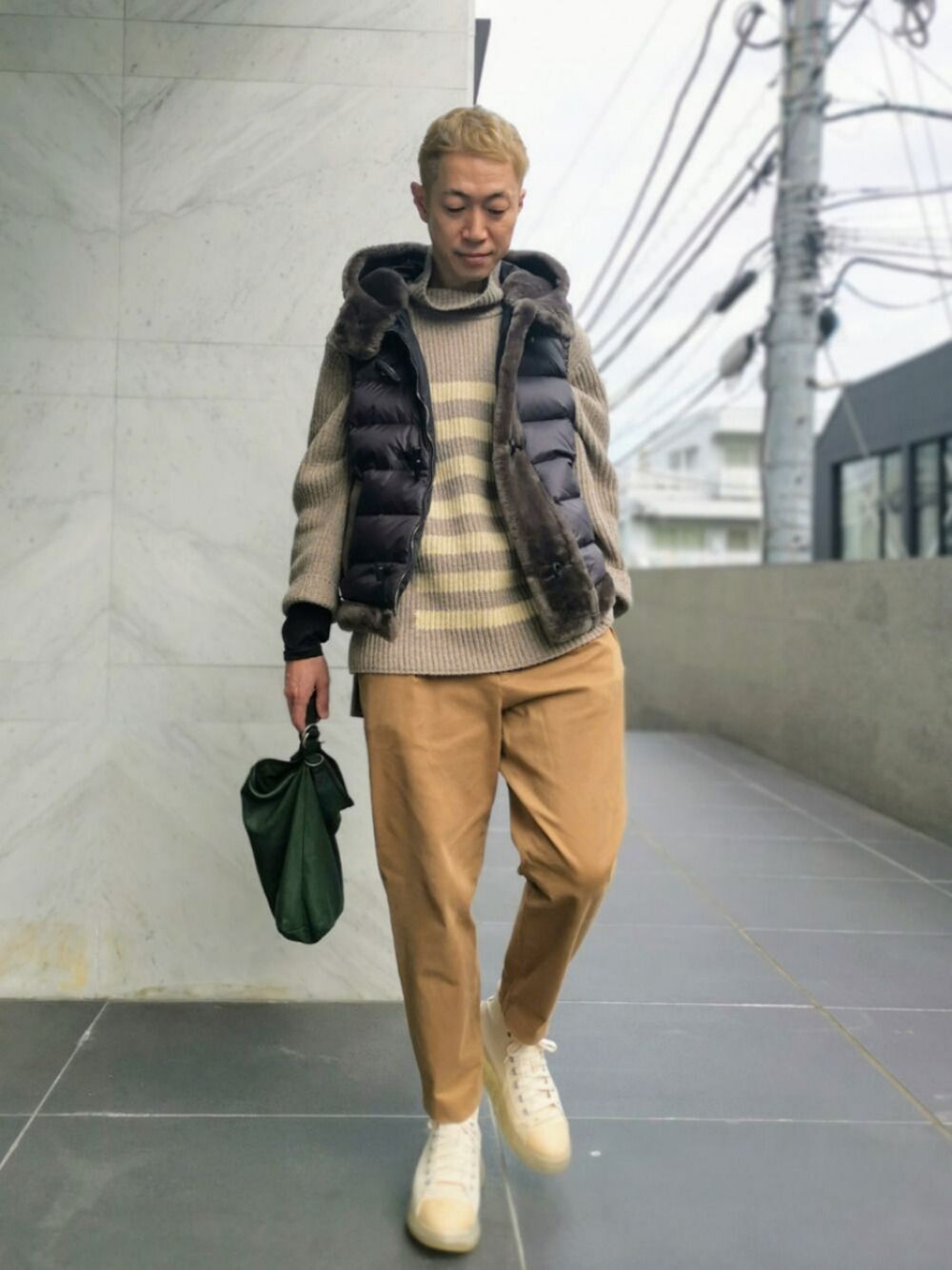 ベージュ系のニット/セーター、ブラック系のダウンベスト、グリーン系のハンドバッグを着用したメンズの秋コーディネートの1枚目の写真